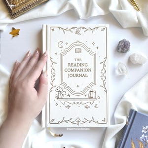 May include: A cream-coloured journal with the title "The Reading Companion Journal" and gold illustrations of flowers and celestial bodies. A gold pen, framed quote, and crystals are also visible. The journal has a cream-coloured ribbon bookmark.