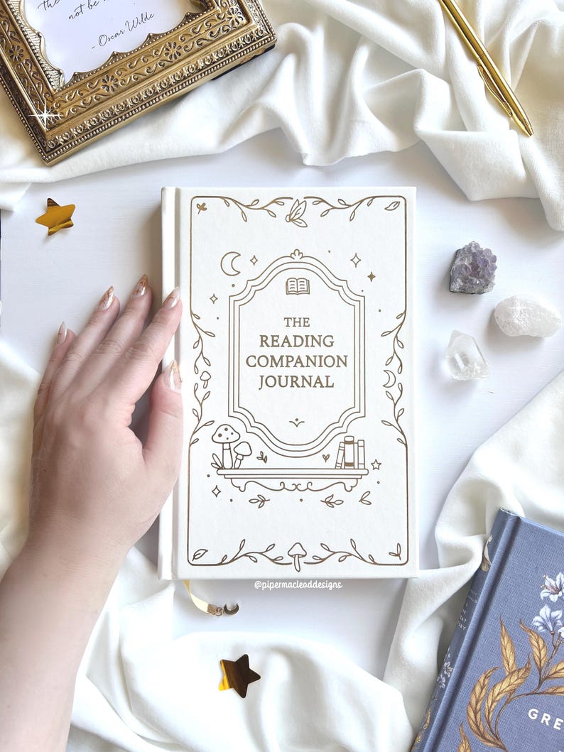 May include: A cream-coloured journal with the title "The Reading Companion Journal" and gold illustrations of flowers and celestial bodies. A gold pen, framed quote, and crystals are also visible. The journal has a cream-coloured ribbon bookmark.