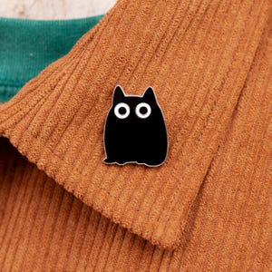 May include: Black enamel pin of a cartoon cat with large eyes. The cat is sitting on a brown corduroy fabric.