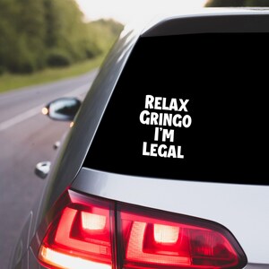 May include: A silver car's rear window features a white vinyl sticker with the words "RELAX GRINGO I'M LEGAL" in a bold font. The car is on a road with green trees visible in the background.