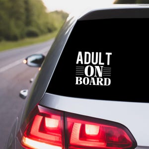 May include: A white vinyl car sticker displaying the words "ADULT ON BOARD" in a bold, sans-serif font. The sticker is on the rear window of a silver car, with a blurred background of a road and trees.