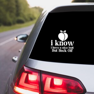 May include: A white vinyl car sticker featuring a peach graphic and the text "I know I have a nice butt But Back Off" is displayed on the rear window of a silver car. The car is on a road.