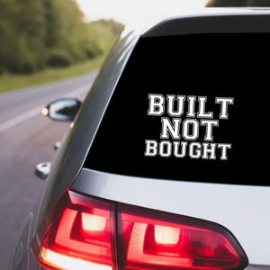 May include: A white vinyl sticker on the rear window of a silver car. The sticker reads "BUILT NOT BOUGHT" in a bold, outlined font. The car is on a road with trees in the background.