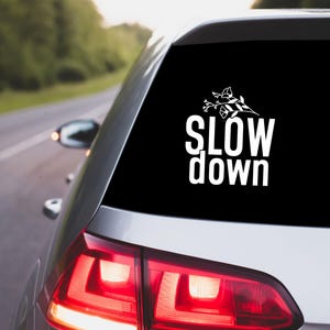 May include: The rear window of a silver car features a black sticker with the white text "SLOW down" and a floral motif. The car is on a road with trees visible in the background.