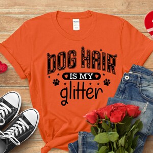 May include: An orange t-shirt with the slogan "DOG HAIR is my glitter" in black lettering. The image includes blue jeans, black trainers, and a bouquet of red roses. The t-shirt has a crew neck and short sleeves.