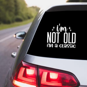 May include: A black car window sticker with the words "I'm not old, I'm a classic" in white. The sticker is on the rear window of a silver car, with a blurred background of a road and trees.