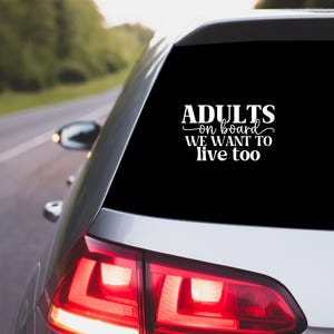 May include: A silver car's rear window features a white vinyl decal with the text "ADULTS on board WE WANT to live too". The car is driving on a road with green trees in the background.