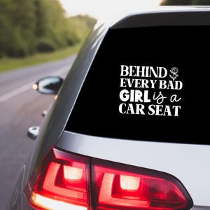 May include: A black car window sticker with white text. The text reads "BEHIND EVERY BAD GIRL is a CAR SEAT." The sticker is on the rear window of a silver car, with a road and trees visible in the background.