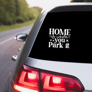 May include: A white decal on a car window with the text "HOME is where you Park it" in a decorative font. The car is silver and driving on a road with green trees in the background.