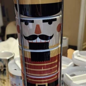 以下が含まれることがあります： 金属製のホルダーに入った、積み重ね可能なセラミック製くるみ割り人形マグセット。マグカップは、くるみ割り人形の顔と、黒、赤、金、青のユニフォームのデザインで装飾されています。セットはシルバーの金属製スタンドで保持されています。