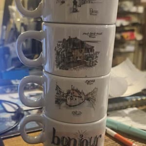 Può includere: Una pila di quattro tazze da caffè in ceramica bianca. Ogni tazza presenta una diversa illustrazione in bianco e nero di una scena urbana europea. La tazza inferiore ha la parola "bonjour".