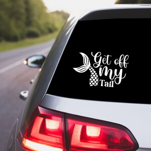 May include: A white vinyl car sticker featuring the phrase "Get off my Tail" and a mermaid tail design. The sticker is affixed to the rear window of a silver car, with a blurred backdrop of a road and trees.
