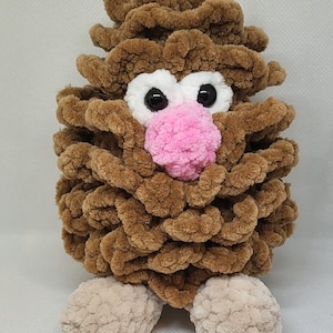 May include: A brown crocheted plush toy with a pink nose, white eyes, and black pupils. The toy has two beige crocheted feet.