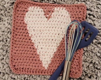 May include: A pink crocheted dishcloth with a white heart design. The dishcloth is lying on a gray fuzzy surface with a blue and silver whisk and spatula on top.