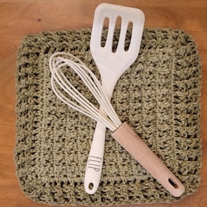 May include: A white kitchen utensil set with a whisk and a spatula. The utensils are on top of a green crocheted dishcloth. The spatula has the word "FLIP" printed on it.
