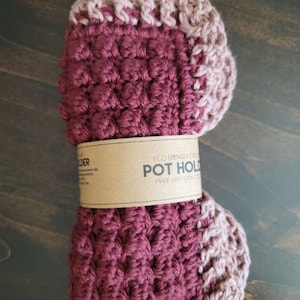 May include: A burgundy and pink crocheted pot holder with a brown label that reads "Eco Friendly Crochet Pot Holder Made with 100% Cotton."