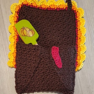 May include: A brown crocheted turkey pot holder with a yellow, orange and red scalloped border. A green spatula with a pumpkin design is tucked into the pocket.