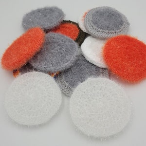 May include: A pile of crocheted dishcloths in white, gray, and orange. The dishcloths are made with a fluffy yarn and have a textured surface.