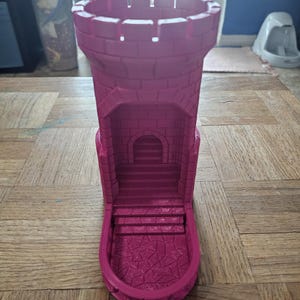 May include: A magenta-colored castle tower dice holder. The tower features a crenelated top, a detailed facade with a small arched doorway, and a set of stairs leading down to a textured base. The design is reminiscent of a medieval fortress.