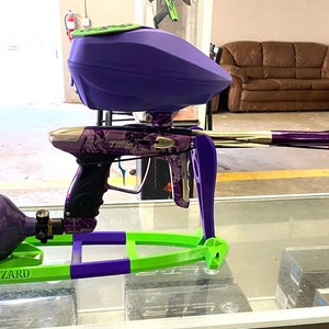 May include: A paintball gun with a purple and gold body, a purple hopper, and a pink and purple tank. The gun has a green and purple frame and the text "B*TCH PLEASE" on the barrel.