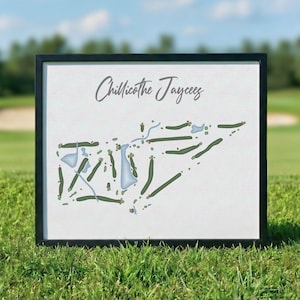 May include: A framed print of a golf course map, featuring green fairways and blue water hazards. The text "Chillicothe Jaycees" is written in cursive at the top. The black frame sits on green grass, with a golf course in the background.