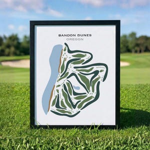 Może przedstawiać: Oprawiony wydruk przedstawiający stylizowaną mapę pola golfowego Bandon Dunes w Oregonie. Grafika wykorzystuje odcienie zieleni, jasnego błękitu i beżu na białym tle, z napisem BANDON DUNES OREGON wydrukowanym w kolorze czarnym i szarym.
