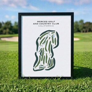 May include: A framed print of the Merced Golf and Country Club in California. The print features a green and white golf course layout with the course name and location printed at the top. The frame is black, and the print is displayed on a grassy surface.