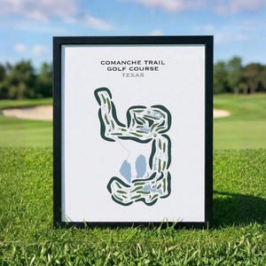 May include: Framed print of the Comanche Trail Golf Course in Texas. The course layout is depicted in green and blue on a white background. The print is in a black frame and is displayed on a grassy surface.