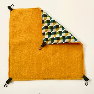 May include: A yellow fleece hamster hammock with a geometric pattern on the inside. The hammock has four black clips to attach it to a cage.