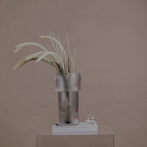 以下が含まれることがあります： リブ模様の透明なガラスの花瓶に、白いドライフラワーが飾られています。花瓶は透明なアクリル製の台座の上に置かれ、その上に本が積み重ねられ、小さな透明なガラスのボウルには白い石が入っています。