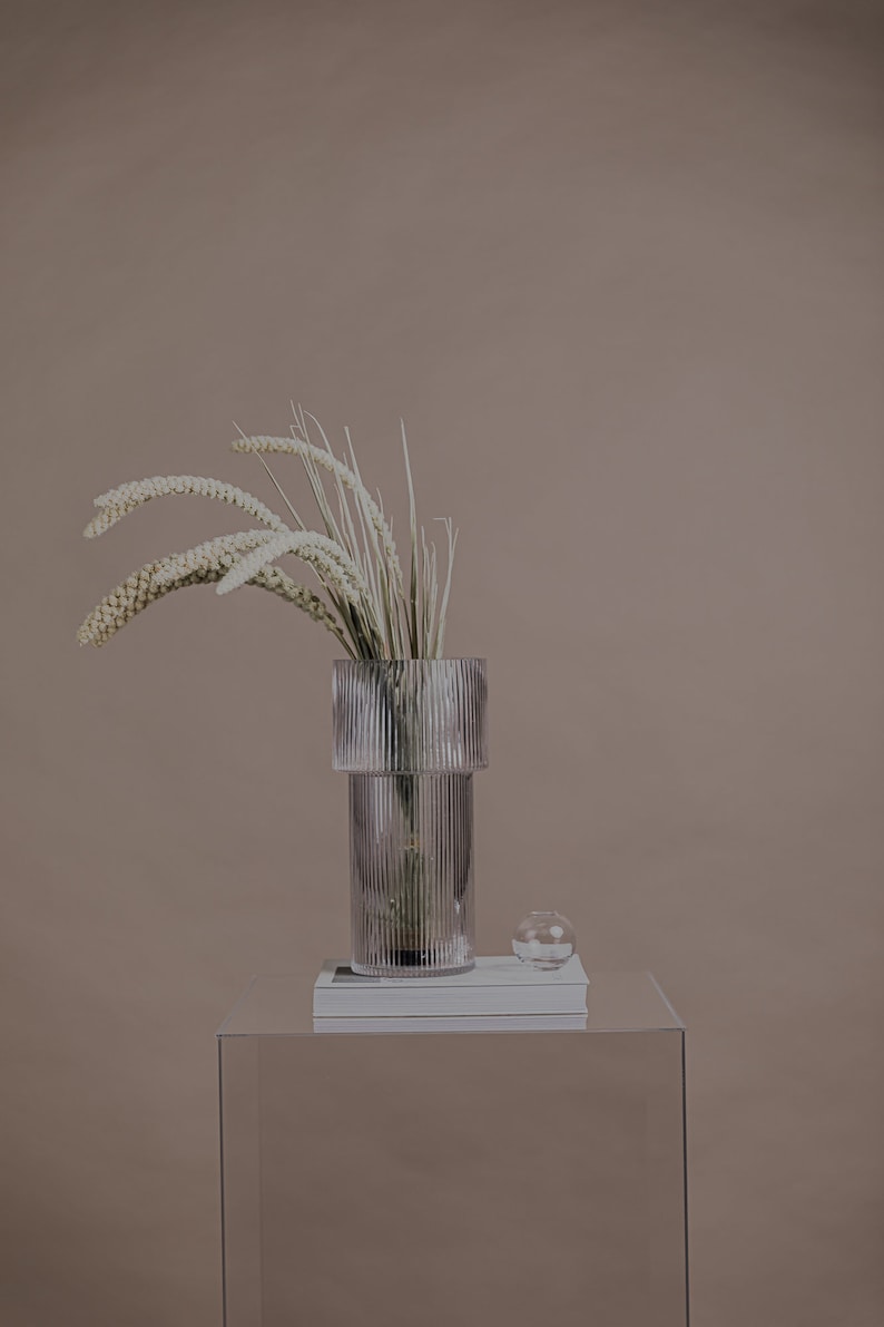 以下が含まれることがあります： リブ模様の透明なガラスの花瓶に、白いドライフラワーが飾られています。花瓶は透明なアクリル製の台座の上に置かれ、その上に本が積み重ねられ、小さな透明なガラスのボウルには白い石が入っています。