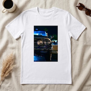 Op de afbeelding: Een wit T-shirt met korte mouwen en een gecentreerde rechthoekige grafische print. De afbeelding toont het vooraanzicht van een donkergekleurde oldtimer in de nacht, met de nadruk op de dubbele ronde koplampen, chromen grille en blauwe verlichting.