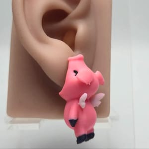 Pode incluir: Um brinco de porquinho rosa com pequenas asas brancas nas costas, detalhes pretos nos olhos e pontas pretas nos pés. O porco é retratado em pé com o focinho voltado para a frente, apresentando um acabamento fosco e liso em toda a peça.