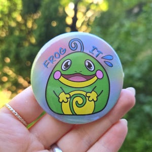 Könnte beinhalten: Ein runder Button mit einem Cartoon-Frosch-Design. Der Frosch ist grün mit rosa Wangen und einem gelben Bauch. Der Button trägt die Worte "FROG IT" in Blau, vor einem blau-rosa Farbverlauf.