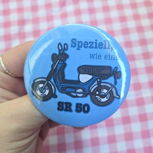 Könnte beinhalten: Ein runder, hellblauer Ansteckbutton mit einer dunkelblauen Illustration eines Mopeds. Der Text lautet Speziell, wie eine SR 50 in dunkelblauer Schrift. Das Design zeigt ein detailliertes Seitenprofil des Rollers mit schwarzen Reifen und Sitz.