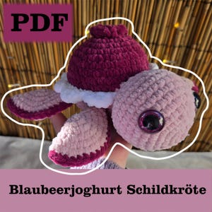 May include: A crocheted turtle amigurumi toy with a pink and purple shell and white accents. The toy has large black eyes and a pink and purple body. The text "Blaubeerjogghurt Schildkröte" is visible below the toy.