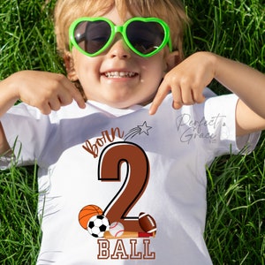 May include: White t-shirt with brown text that says "born 2 ball" with a brown number 2 and a star. The number 2 is surrounded by a baseball, basketball, soccer ball, and football.