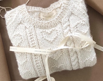 May include: A white knitted sweater with a cable knit pattern. The sweater is folded and tied with a white ribbon that has the text "DREAMLAND KNITS" printed on it.
