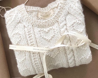May include: A white knitted sweater with a cable knit pattern. The sweater is folded and tied with a white ribbon that has the text "DREAMLAND KNITS" printed on it.
