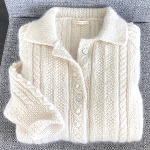 Puede incluir: Cárdigan color crema con botones y cuello. El suéter de punto presenta varios patrones, incluyendo diseños de cable y texturizados. La etiqueta dice "Dreamland Knits".