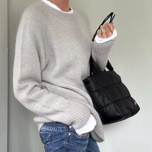 Pode incluir: Um suéter de malha cinza claro com gola redonda e mangas compridas. O suéter é combinado com jeans azuis e uma sacola tote acolchoada preta. Uma camisa branca é visível por baixo do suéter.