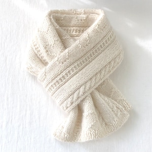 Peut inclure: Une écharpe en tricot de couleur crème avec un motif en câble. L'écharpe est pliée en deux et nouée.