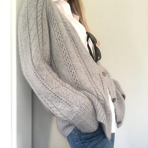Könnte beinhalten: Ein hellgrauer Strickcardigan mit einem Zopfmuster und einem V-Ausschnitt. Der Cardigan hat einen Knopfverschluss und wird über einem weißen Hemd getragen.