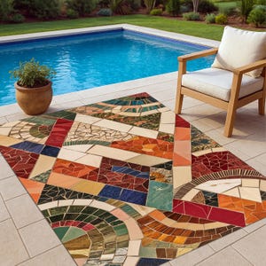 Op de afbeelding: Een kleurrijk buitenkleed met een mozaïektegelontwerp. Het kleed heeft geometrische patronen in tinten rood, oranje, blauw, groen en bruin. Een houten stoel met een wit kussen staat in de buurt, naast een zwembad.