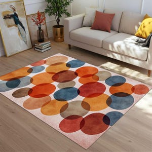 May include: A rectangular rug featuring an abstract pattern of overlapping circles in shades of orange, red, blue, and brown. The rug is set on a wooden floor, with a cream-colored backdrop. The rug is a decorative element for a living room.