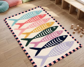 May include: A rectangular area rug with a cream-colored ribbed background featuring seven stylized fish in light blue, red, pink, yellow, dark green, dark blue, and light purple. The rug has a border with alternating pink and dark blue squares.