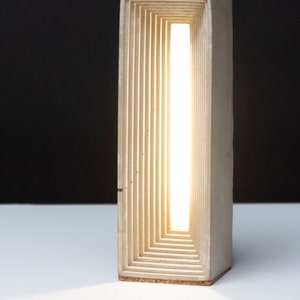 K&ouml;nnte beinhalten: Eine moderne Tischlampe aus Beton mit einem Licht, das durch eine Reihe von &uuml;bereinanderliegenden rechteckigen &Ouml;ffnungen scheint.