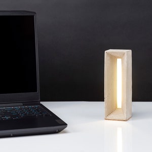 K&ouml;nnte beinhalten: Eine moderne Betonlampe mit einem warmwei&szlig;en Licht, das durch eine Reihe von rechteckigen &Ouml;ffnungen scheint. Die Lampe steht auf einer wei&szlig;en Oberfl&auml;che neben einem schwarzen Laptop.