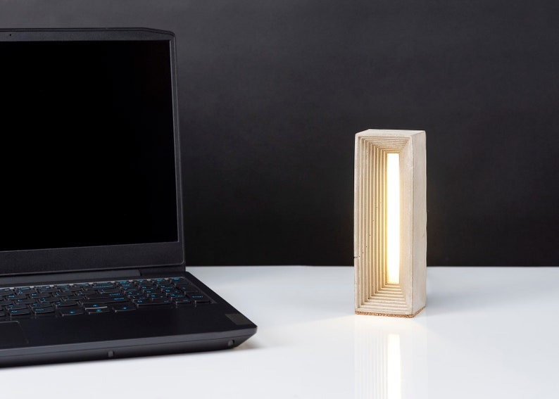 K&ouml;nnte beinhalten: Eine moderne Betonlampe mit einem warmwei&szlig;en Licht, das durch eine Reihe von rechteckigen &Ouml;ffnungen scheint. Die Lampe steht auf einer wei&szlig;en Oberfl&auml;che neben einem schwarzen Laptop.