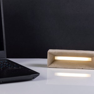 K&ouml;nnte beinhalten: Eine moderne Betonlampe mit rechteckiger Form und einem d&uuml;nnen, wei&szlig;en Lichtstreifen im Inneren. Die Lampe steht auf einer wei&szlig;en Oberfl&auml;che neben einem schwarzen Laptop.